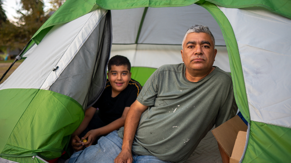 An older man and a young boy are looking out from the entrance of their tent.