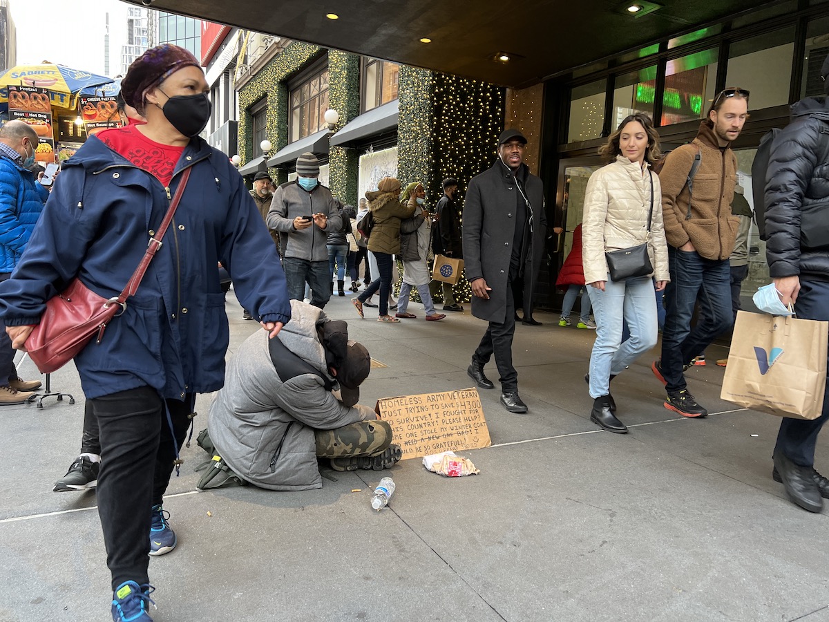 People walk around a homeless person with a sign on a sidewalk