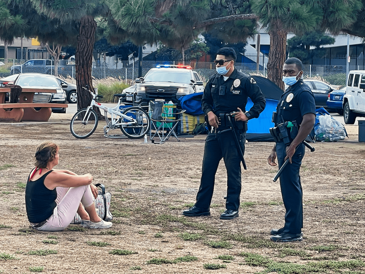 Two police officers talk to a homeless woman sitting down in an empty lot.