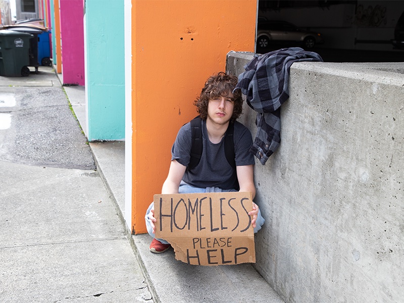 A young man holds a sign that reads 'homeless please help'.