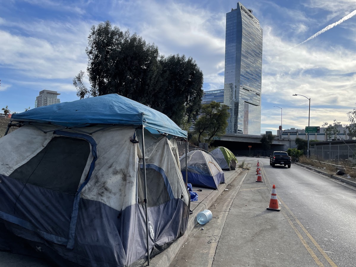 A line of tents can be seen on the sidewalk, with a skyscraper visible in the background.