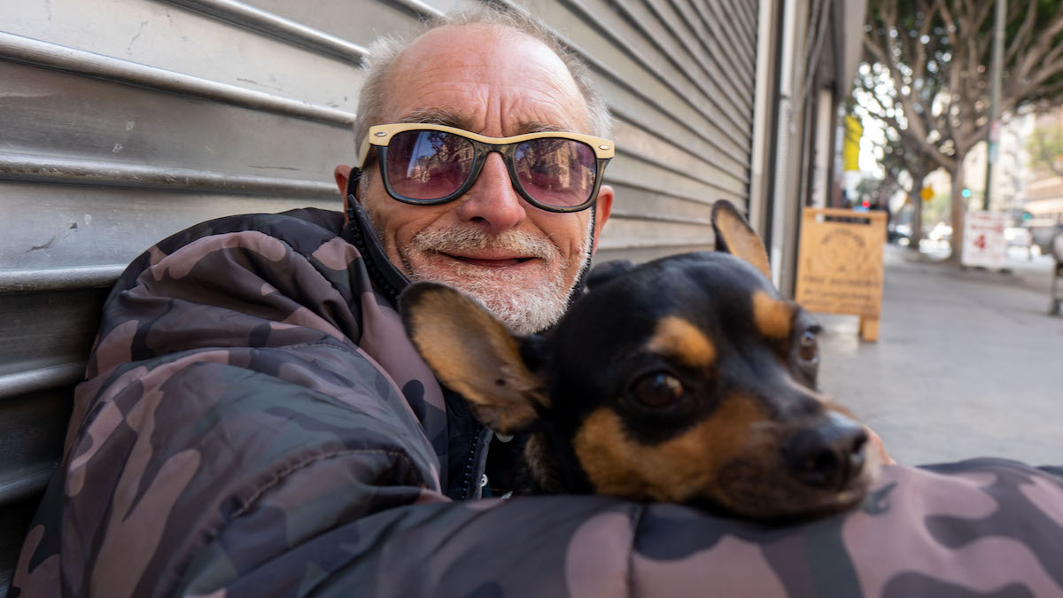 Homeless man wearing sunglasses hugging a dog outside on the sidewalk.
