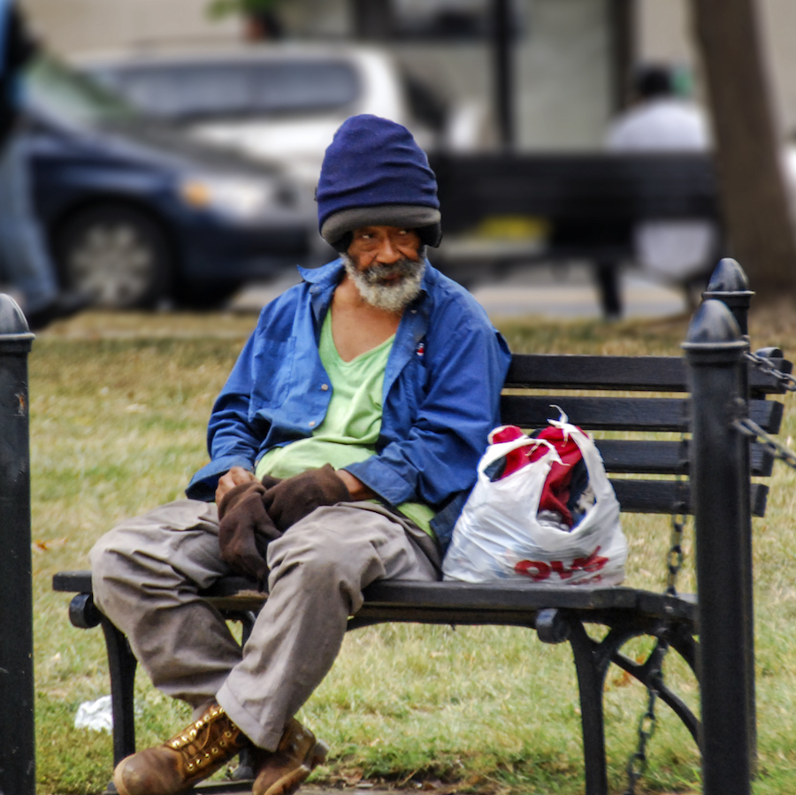 A homeless man wearing a beanie sits on a bench in a park.