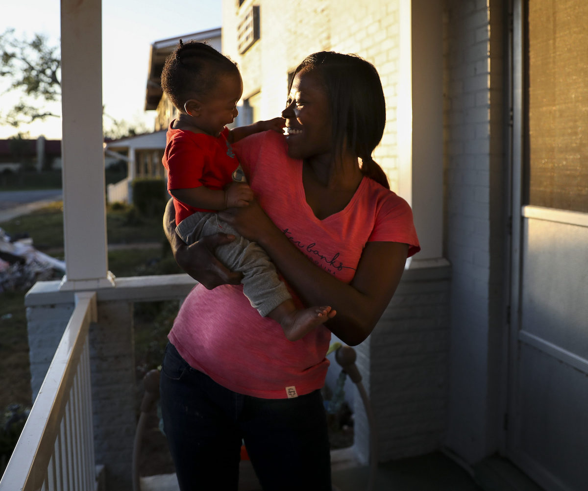 A Black mother holds her baby on the porch of a house at sunset.