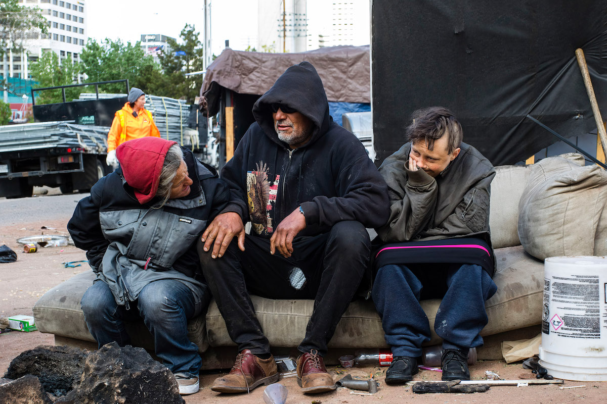 Three homeless men sit on a couch outdoors, talking to each other.