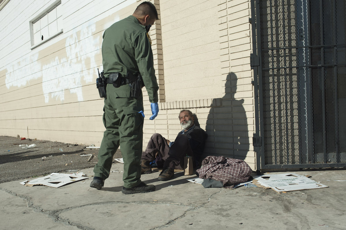 A police officer stands over a homeless man sitting against a wall.