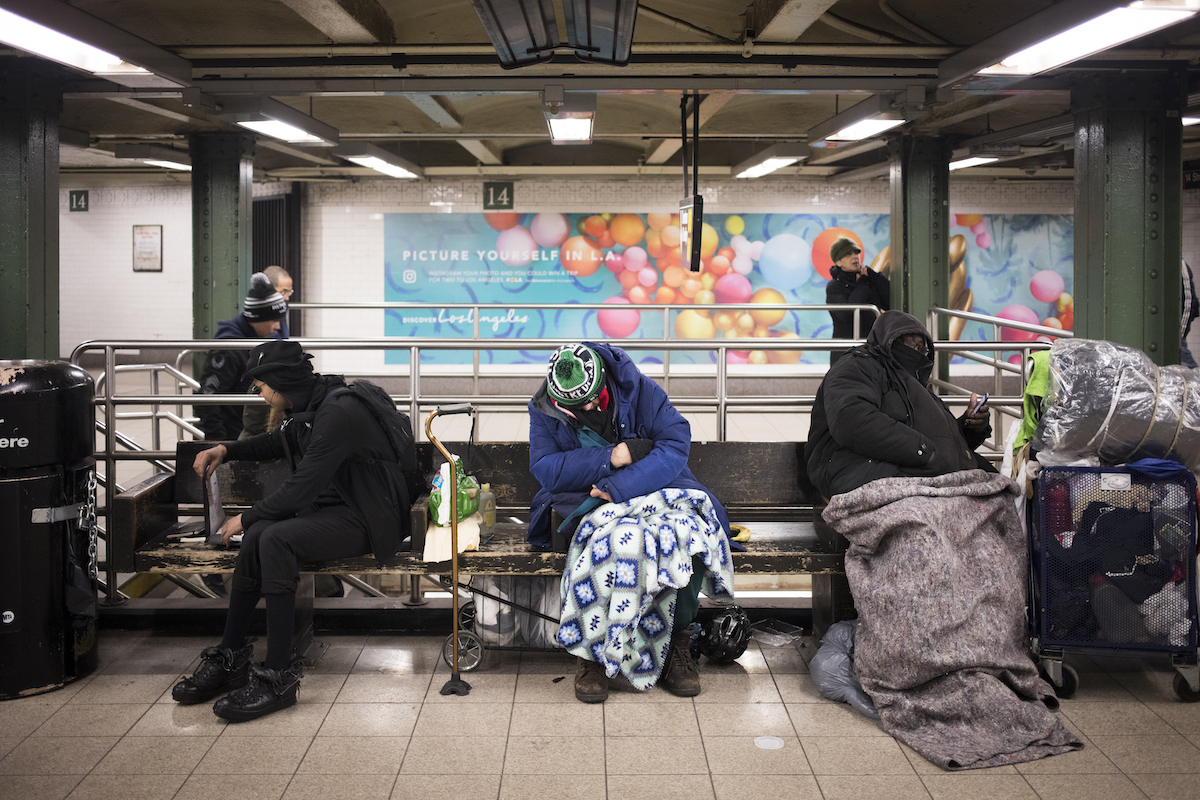 Three homeless people sit on a bench inside a subway station.