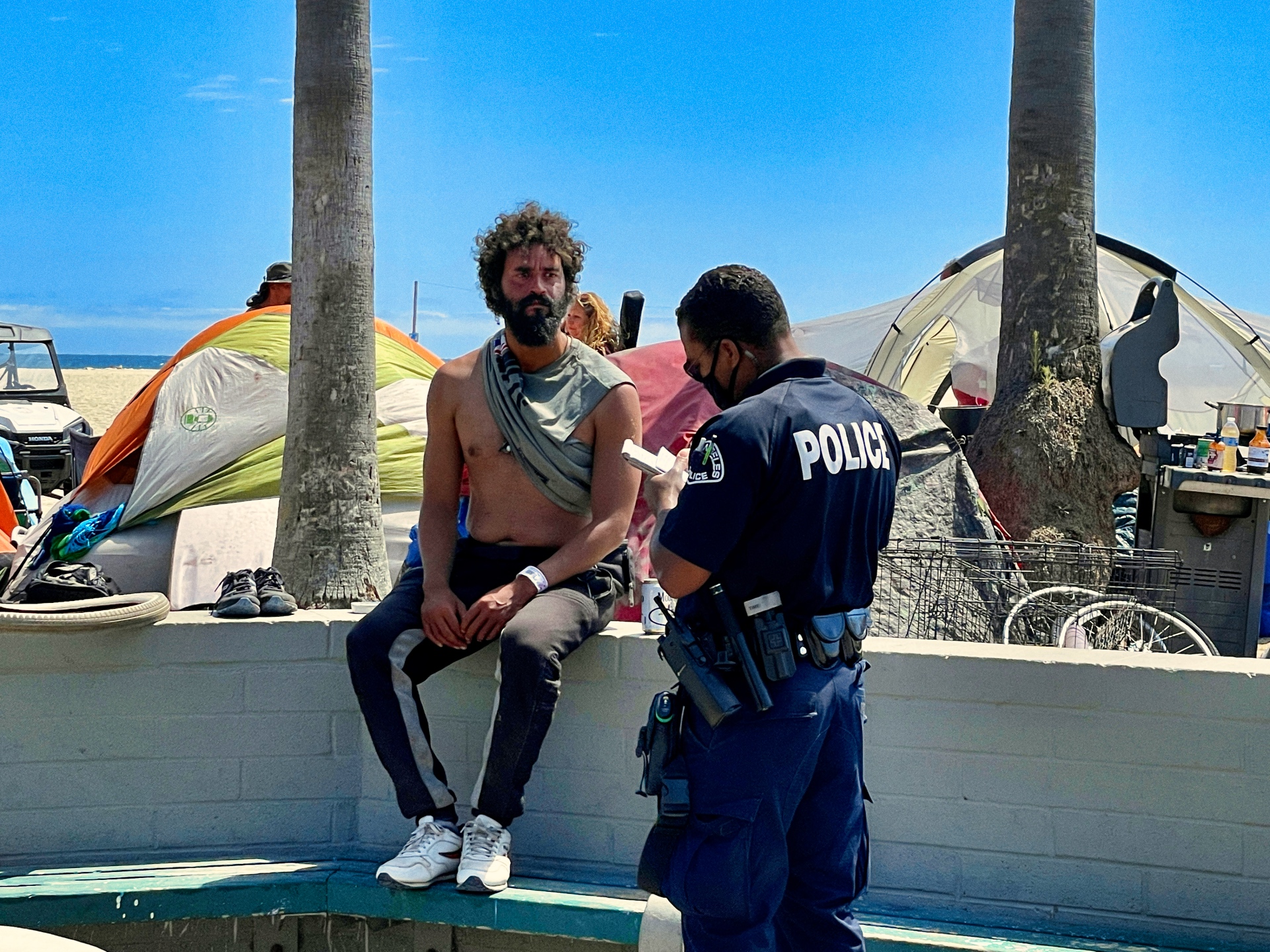 A man not wearing a shirt sits on a short wall while a police officer writes him a ticket.