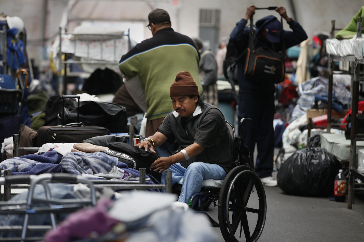 A man in a wheelchair arranges his bag and other items on a bed in a homeless shelter.