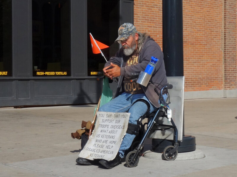 a homeless veteran sits on the street with a sign
