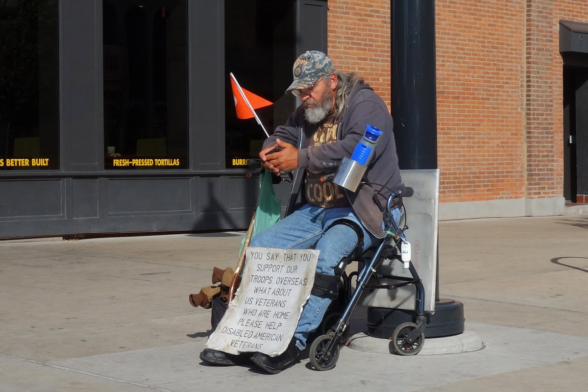 a homeless veteran sits on the street with a sign